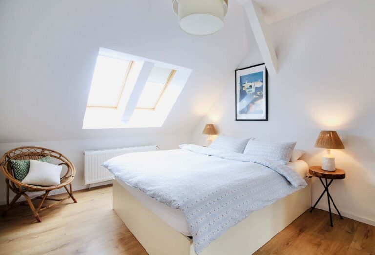Attic bedroom interior with a bed, skylight, and wicker chair. Cozy room with white walls, wood floors, and lamps.