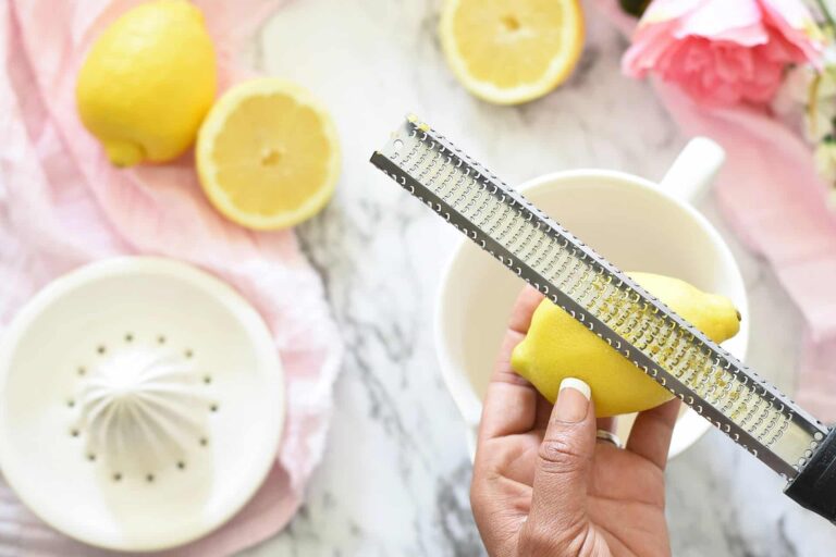 Lemon zest. A hand zests a lemon with a microplane over a white bowl. Lemons, juicer, and flowers in the background.