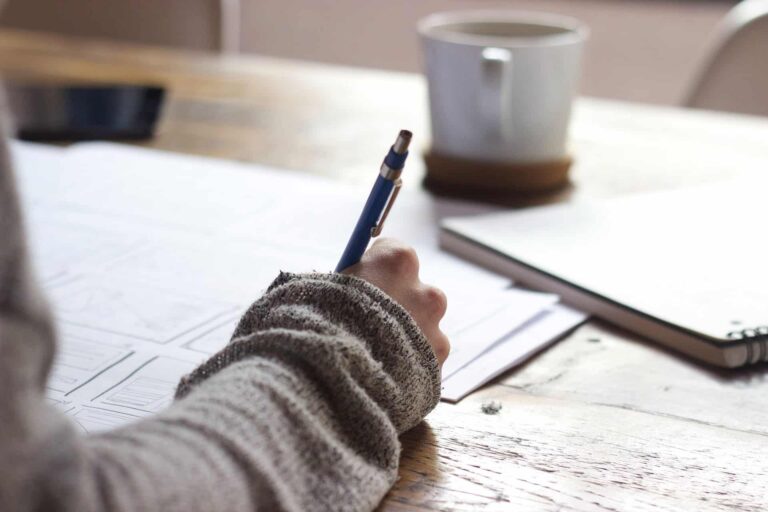 Person writing on paper at desk with pen, notebook, and coffee. Studying, working, or creative writing concept.