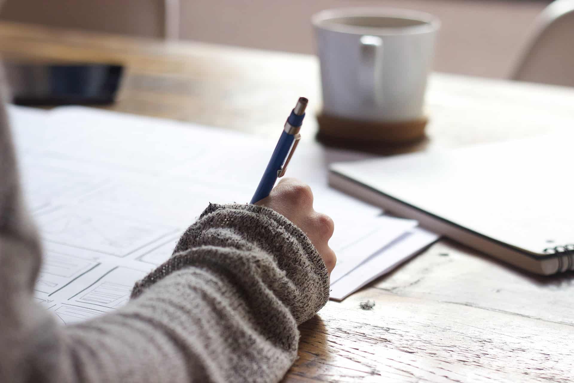 Person writing on paper at desk with pen, notebook, and coffee. Studying, working, or creative writing concept.
